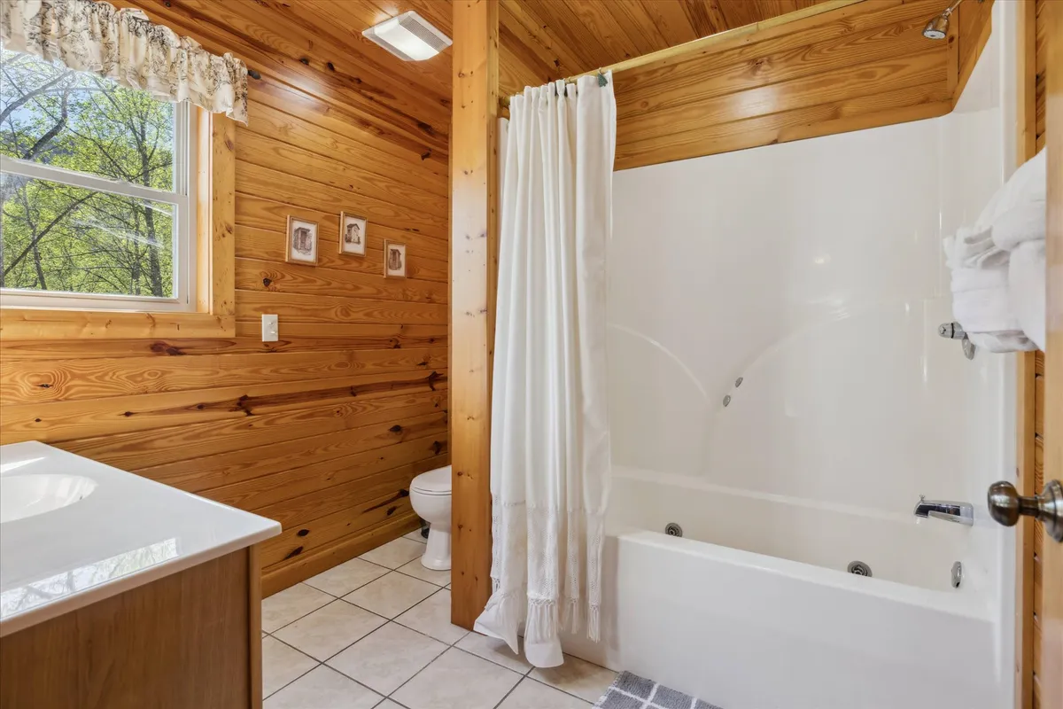 An en suite bathroom featuring a spacious shower area, elegant vanity, wood paneling, and a window with a view of the surrounding woods. The space feels intimate and relaxing, ideal for self-care routines and unwinding.