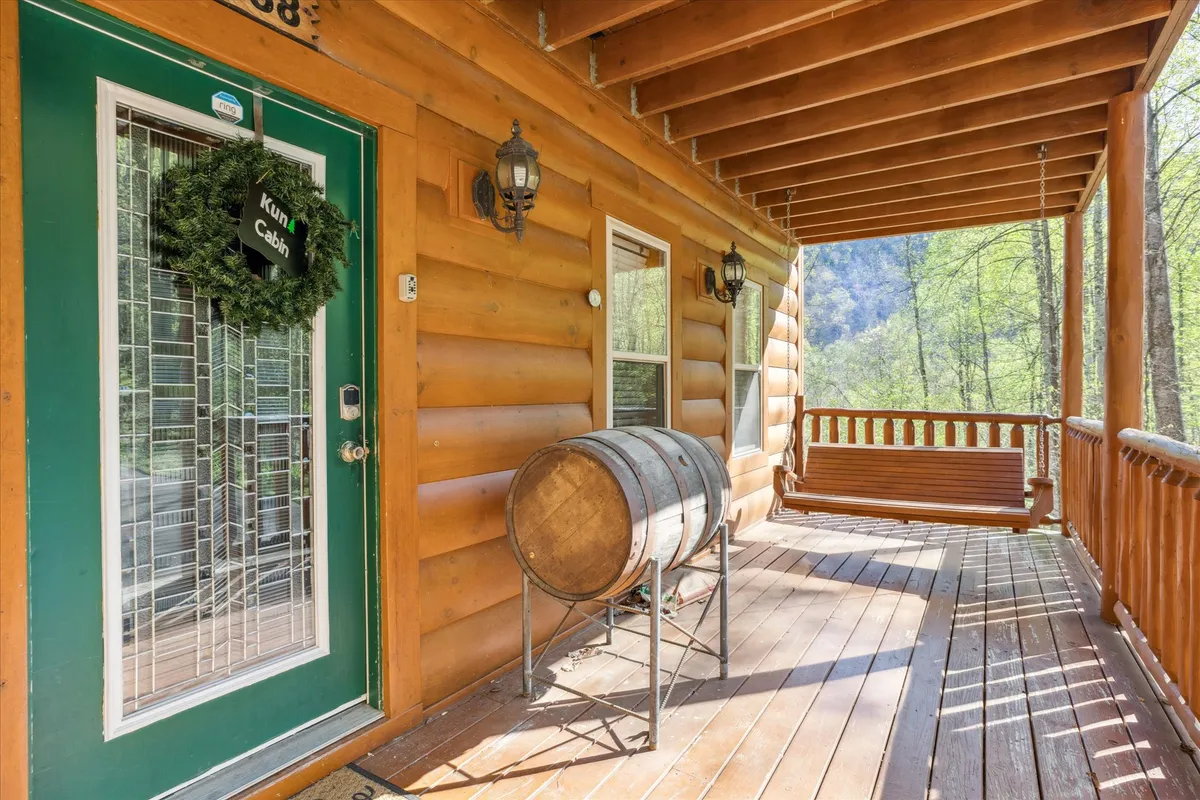 The cabin’s green front door opens onto a cozy wooden porch, complete with a swinging bench and rustic barrel table. Log siding and comfortable seating invite you to linger and take in the serene woodland views, whether at sunrise or dusk.