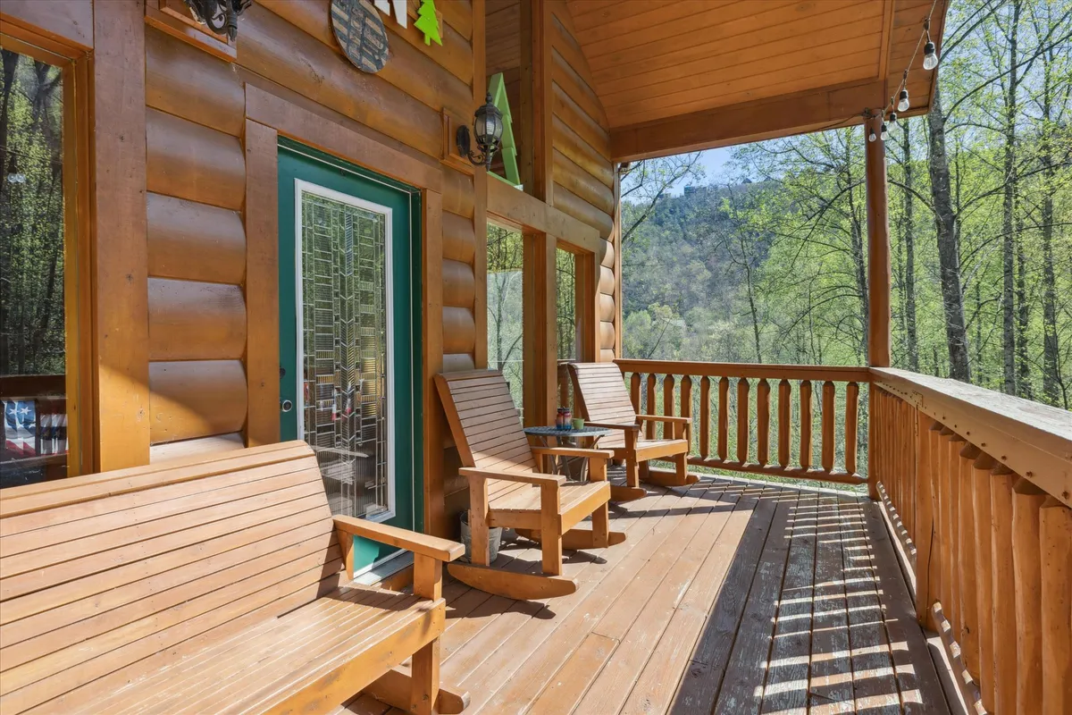 An inviting exterior view featuring wooden rocking chairs on the front porch, a decorative glass front door, and lush surrounding trees. The tranquil setting suggests a peaceful spot for friends to gather, relax, and enjoy nature together, hinting at the cabin’s connection to its beautiful surroundings.