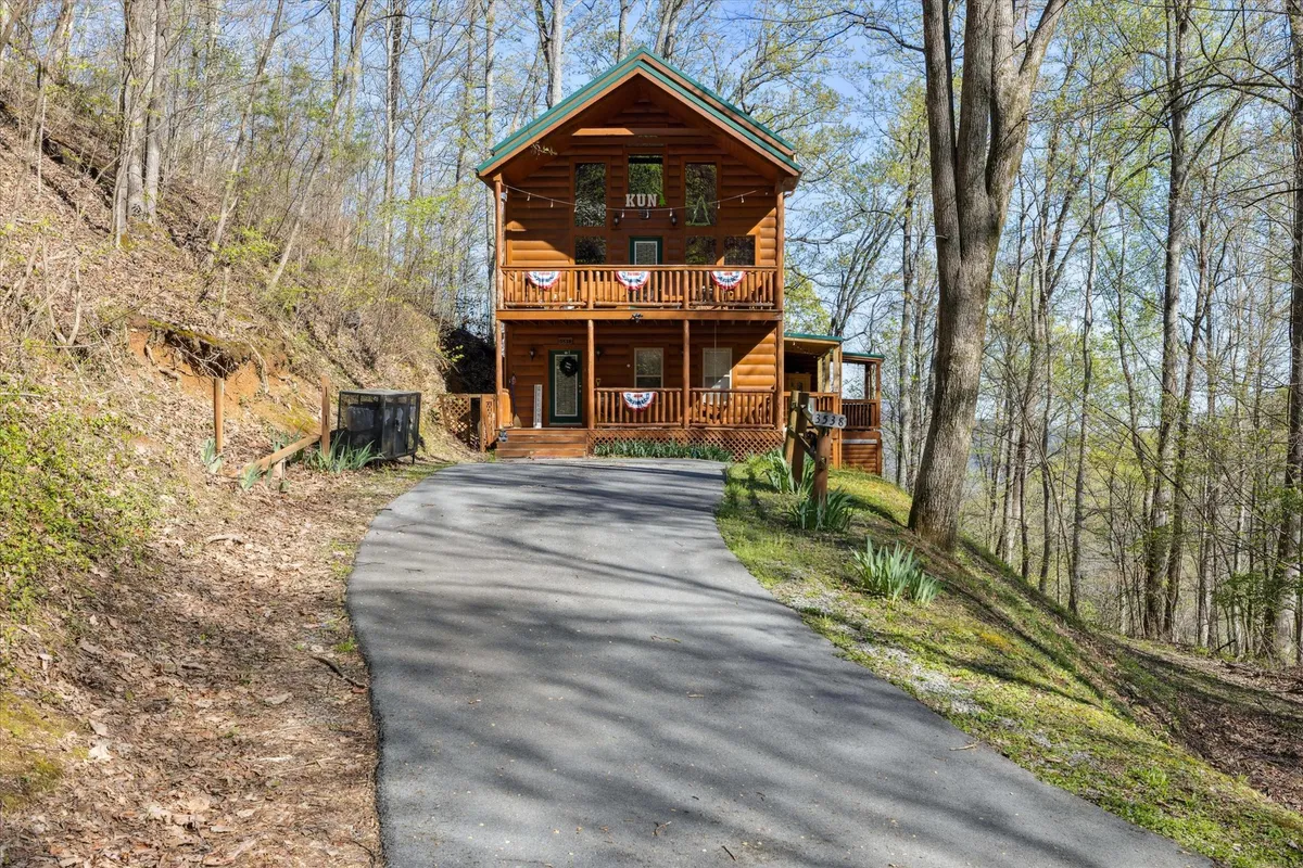 The hero image features a charming log cabin with a distinctive green roof, inviting balconies with rustic railings, and festive decorative bunting. The cabin sits nestled among lush trees, evoking warmth and tranquility, with a spacious porch perfect for watching sunrises or relaxing outdoors. The yard offers space for evening campfires, while the serene natural surroundings promise peace and privacy.