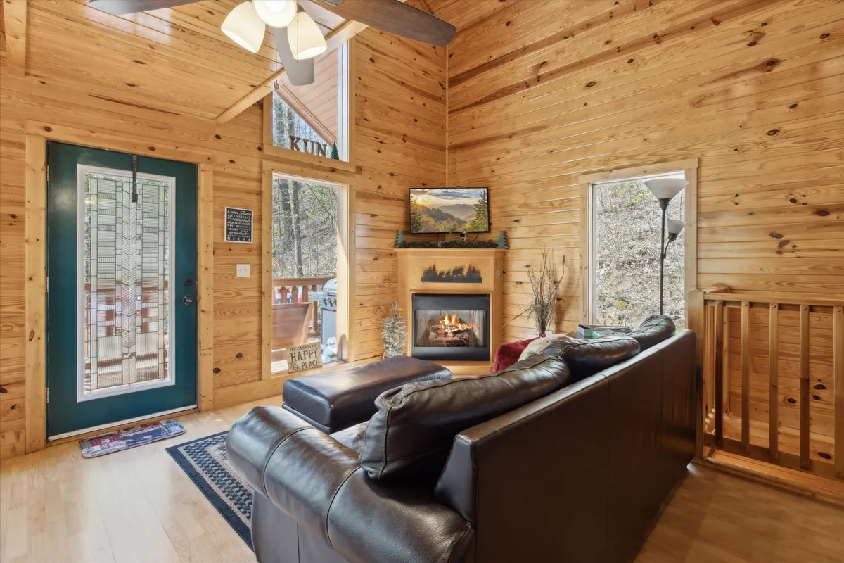 This image presents the heart of the living room, highlighting a leather sofa arranged around a crackling fireplace with large windows framing tranquil woodland views. Warm wooden walls and decorative artwork add to the inviting, homey atmosphere, making the space ideal for relaxation and socializing.