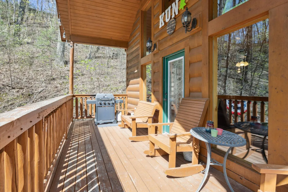 A spacious wooden deck outfitted with rocking chairs, a grill, and a small table provides a casual outdoor living space surrounded by trees. This inviting area is set up for family barbecues, stargazing, and sharing meals outdoors after exploring the Smokies.