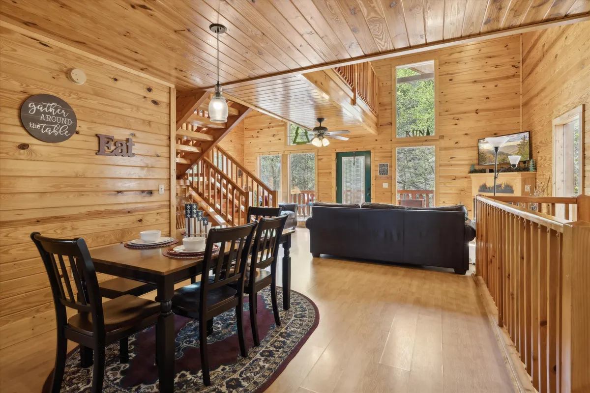 This image showcases the dining area, featuring a large wooden table and chairs set against the backdrop of a cozy living space with sofas, stairs, and generous windows. The open layout invites shared meals and easy flow between social spaces, echoing the section’s focus on connection.